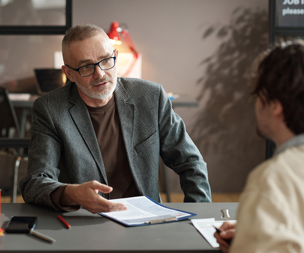 Mature businessman in eyeglasses discussing work experience in resume with candidate during job interview
