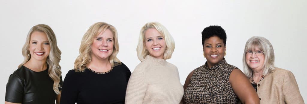 Five women wearing business attire are smiling while standing in a group